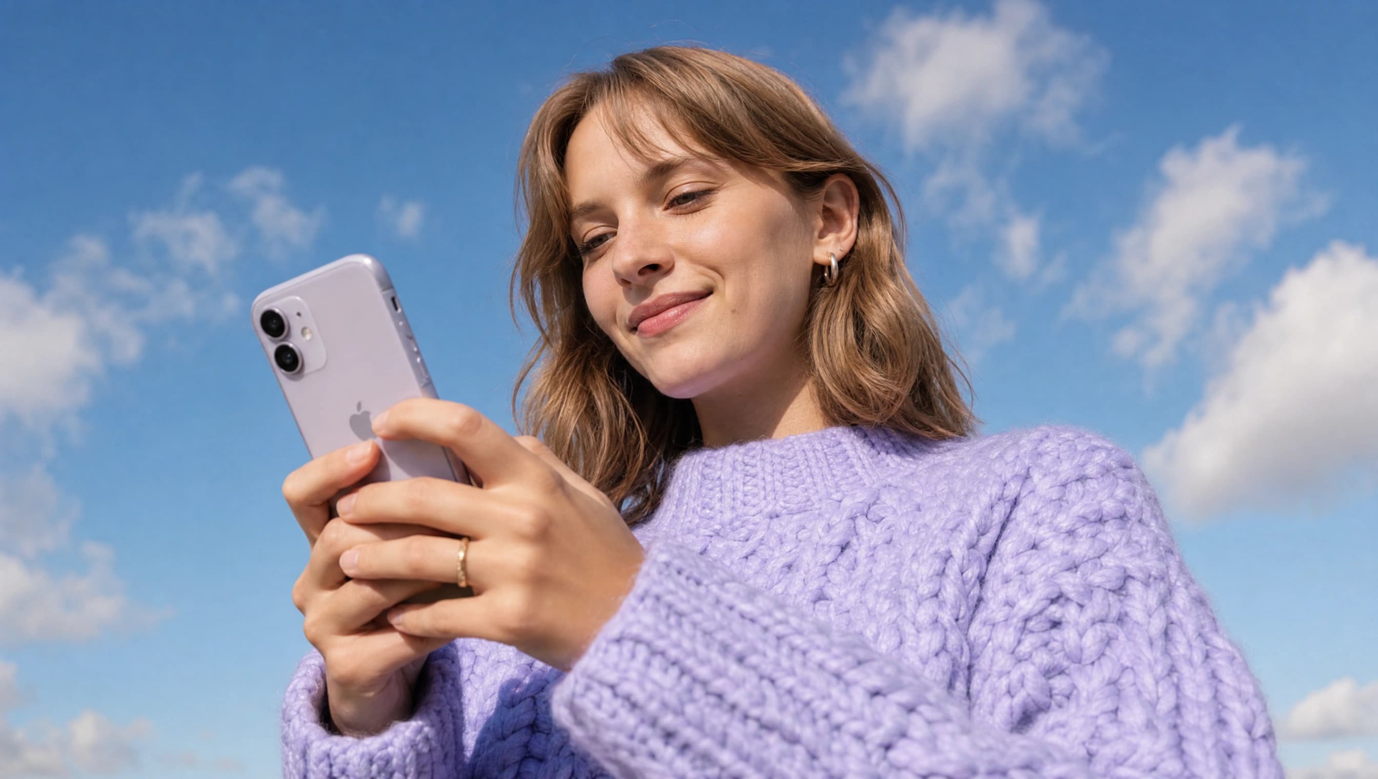 A woman in a lilac knitted jumper smiles as she looks at her phone