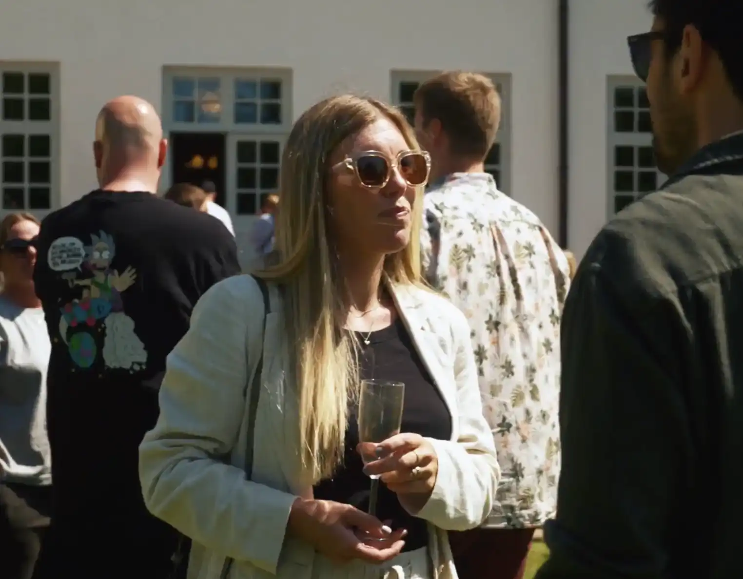 Mennesker samlet udendørs en solskinsdag, forrest en kvinde med solbriller og et glas i hånden