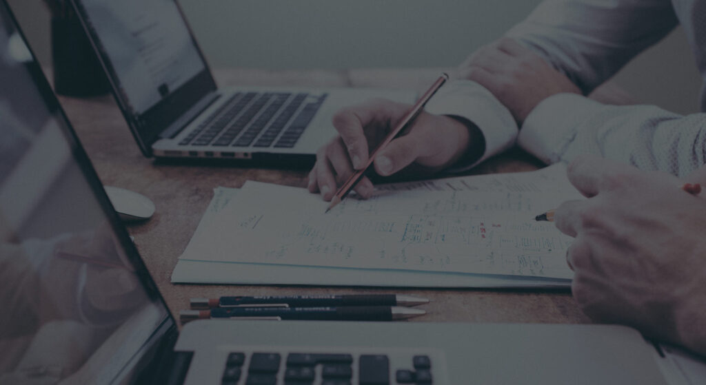 Hænder på skrivebord med papir og computer.